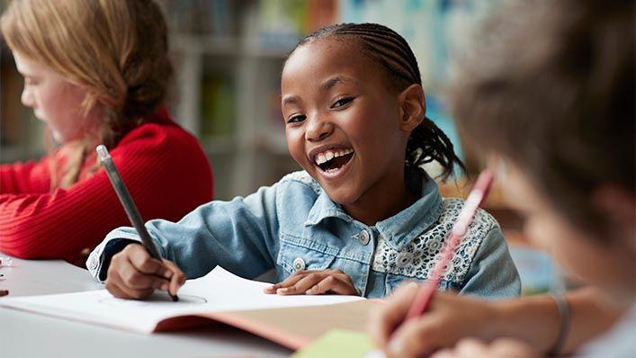 A child in a classroom smiles widely as she works on an assignment