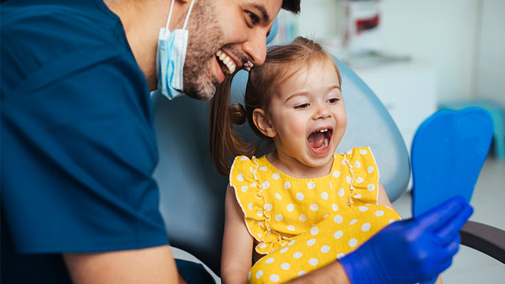 A friendly dentist is smiling and showing a happy child patient her teeth.