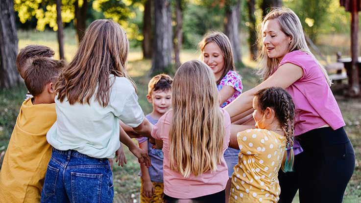 A camp counselor and a group of kids circle up to play a game outside.