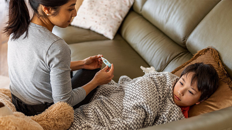 In their living room, a mother holds a thermometer while looking at her young son resting on a couch with a pillow, blanket and teddy bear