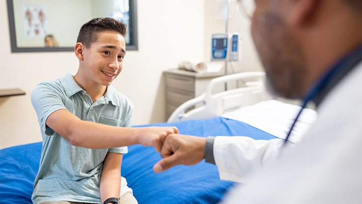 A doctor gives a fist bump to a boy sitting on the edge of an examination table.