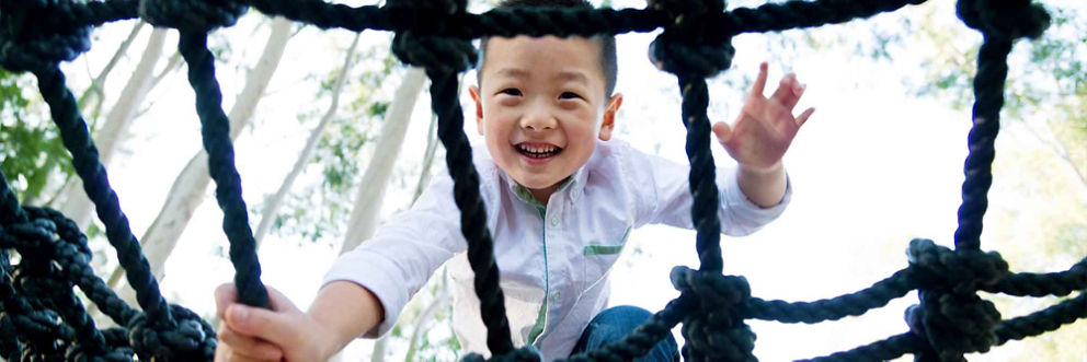 A boy smiles down at the camera while he plays on the rope jungle gym.