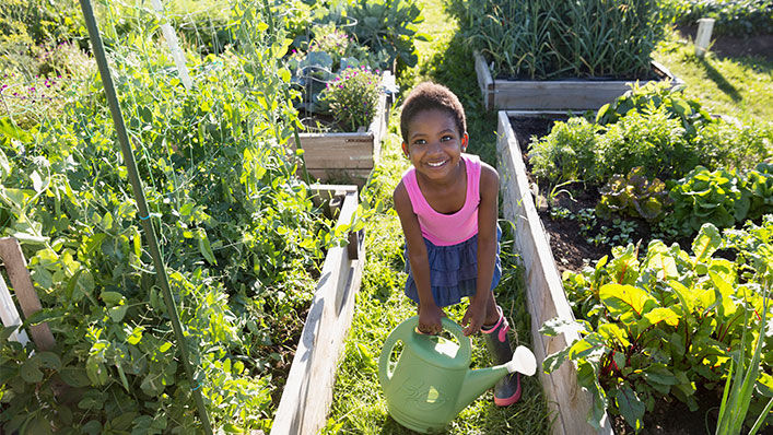 A girl stands smiling between raised garden beds while she holds a watering can.