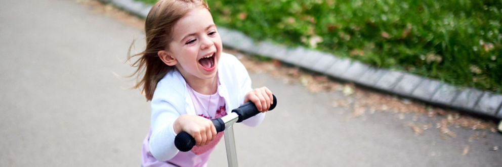 A young girl laughs as she rides on a push scooter outside.