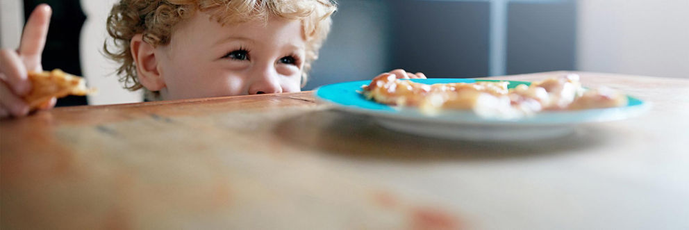 A young boy sneaks a piece of pizza from a plate left on the kitchen table.
