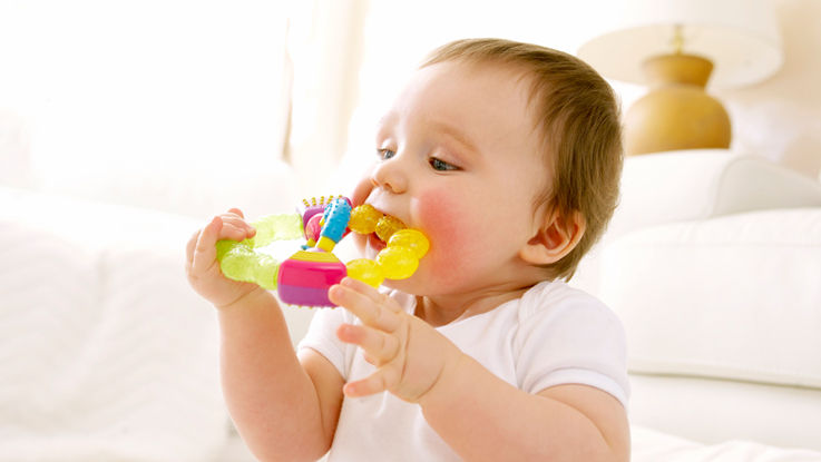 A baby sits in a family room, chewing on a teething ring.