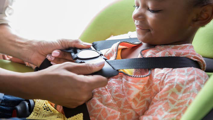 A little girl smiles while her mom buckles her car seat.