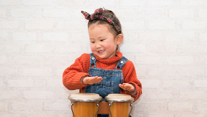 A smiling young girl plays with a set of toy bongos.