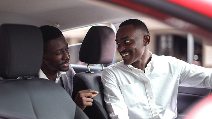 A rideshare driver smiles as he speaks with a client in the backseat of his car