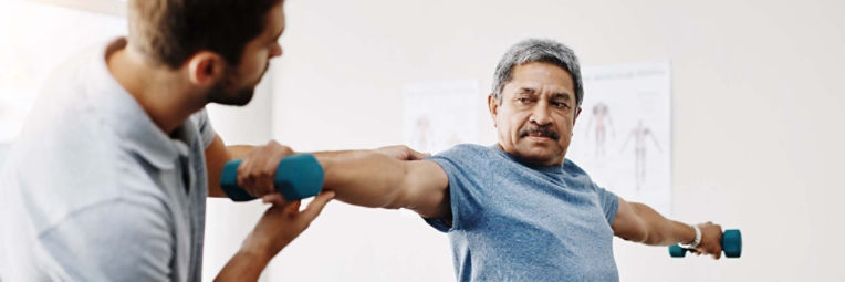 An older man lifts free weights with his arms straight out at his sides under the direction of a physical therapist.