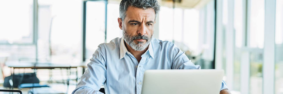 An older man sitting in a sunny room looks intently at his laptop screen