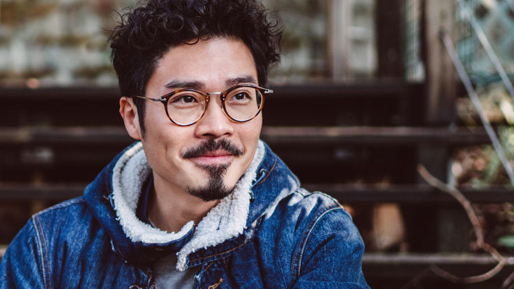 A man with glasses sits on a porch outside as he smiles and looks into the distance.