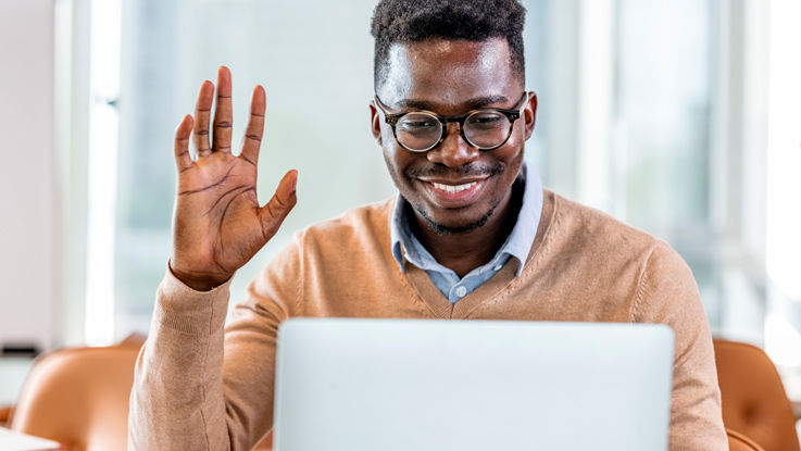 Sitting in a conference room chair, a young man smiles and waves at his laptop screen.