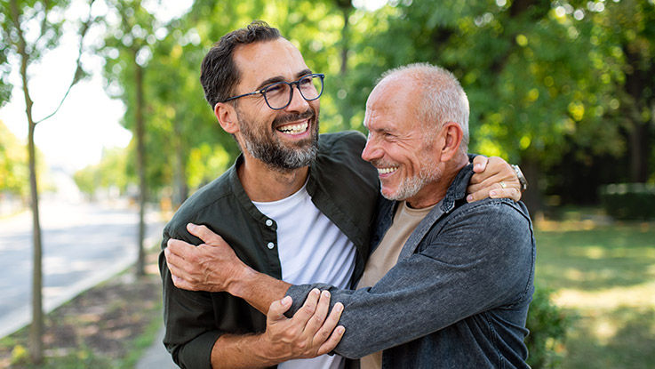 A middle-aged man and an older man hug on a sidewalk.
