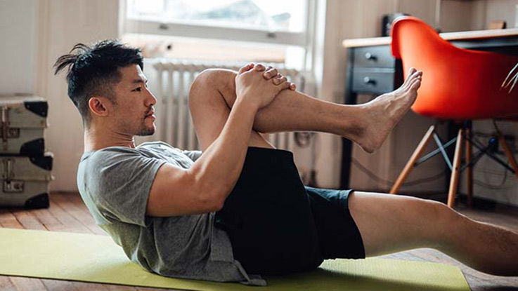 A man lies on his back on a yoga mat in his apartment, grasping his leg to attempt a pose.