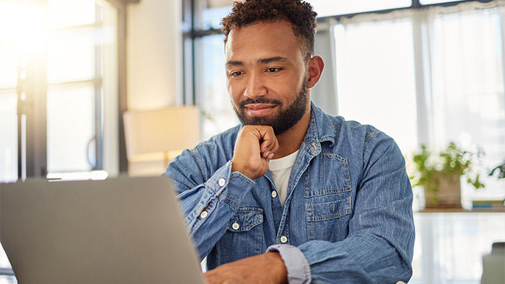 A smiling man rests his chin on his hand while looking at a laptop computer.