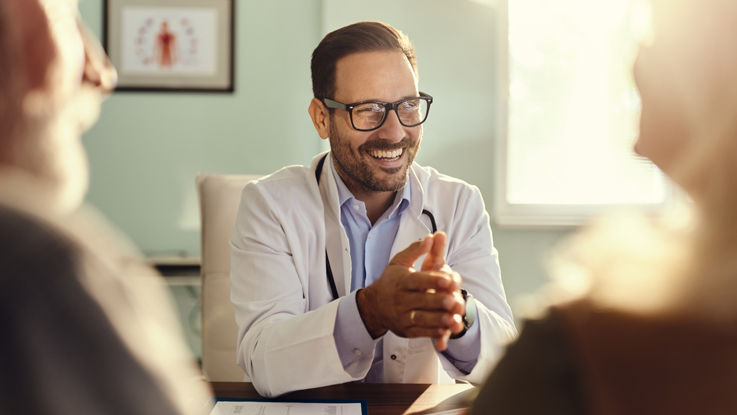 Sitting at his office desk, a doctor smiles as he talks with an older man and woman.