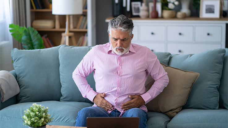 A man presses against the muscles in his lower abdomen while sitting on a couch.