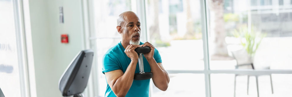 Older man working out in a gym, using a dumbbell and doing squats.