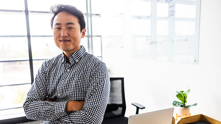 A man confidently looks at the camera as he sits on the desk in his office at work.