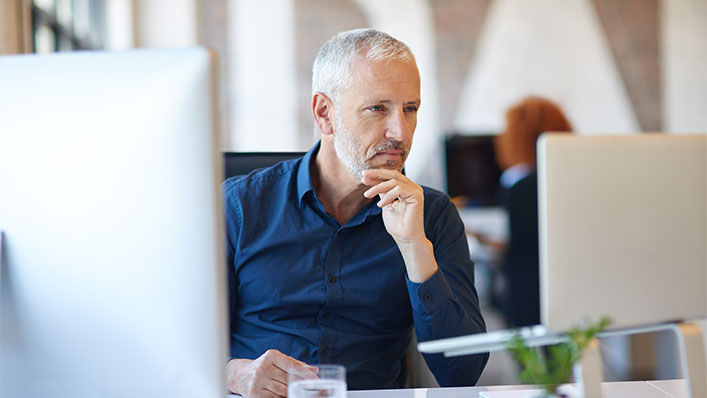 Sitting in an office, a man looks carefully at the laptop screen on his desk.