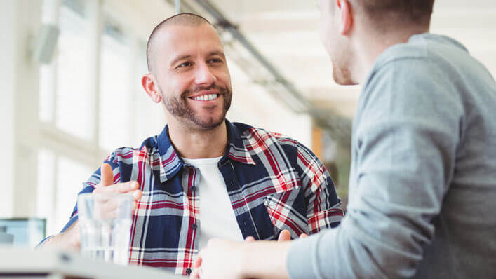 Two men in casual clothing smile as they talk to one another at a meeting.