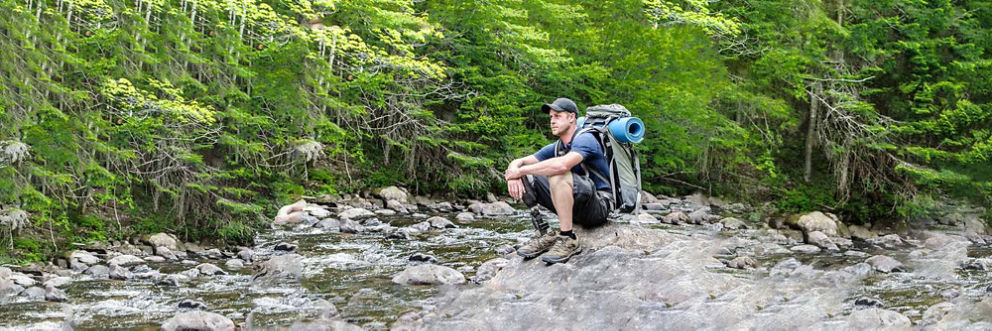  A man with a prosthetic leg sits on a rock in a woodland stream. He wears hiking gear and is carrying a large backpack.