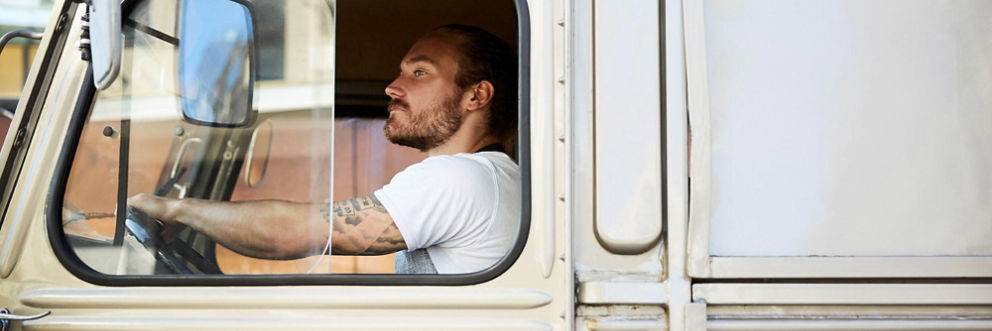 Man in a white t-shirt with a tattoo sleeve driving a truck.
