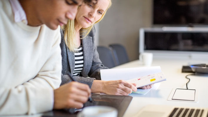 Sitting at a conference room table with a report in her hands, a woman looks at the tablet of the man next to her as he draws on the screen.”