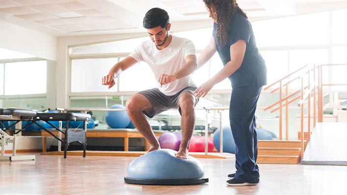 Physical therapist assisting man barefoot trying to balance on a Bosu ball.