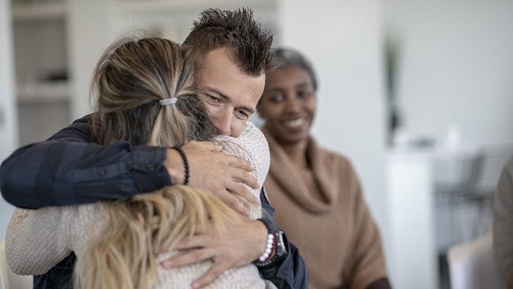 A young caucasian man is hugging a female during a group therapy session. He is hugging her tight and feeling a sense of relief.