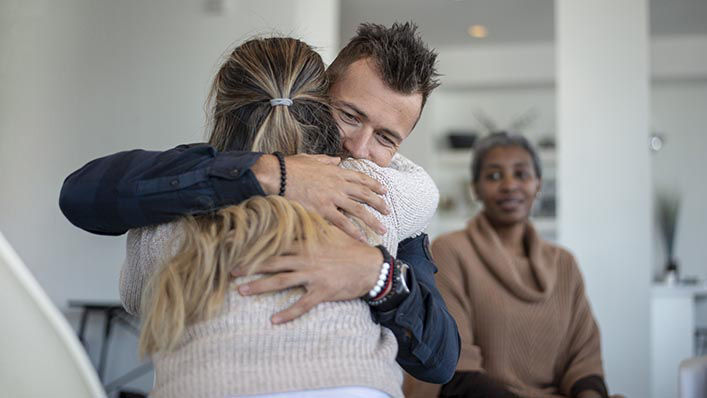 A young caucasian male is hugging a female in a group therapy class. He is hugging her tight and feeling a sense of relief.