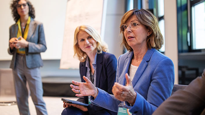 A woman wearing a blazer and glasses gestures as she speaks in a board meeting.