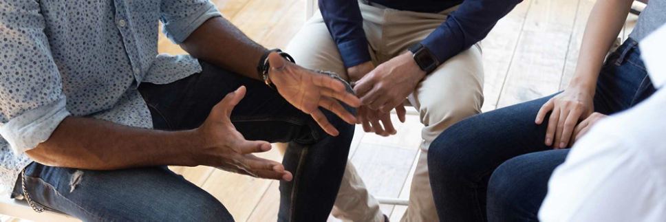 Five people at a support group sit in a close circle of folding chairs and listen as one man speaks and gestures emphatically.