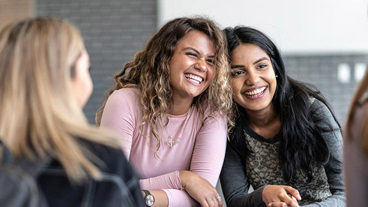 Two women with shiny white teeth smile and laugh as they talk with friends