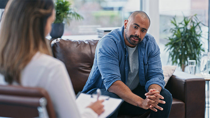  A man sits on a couch with his hands folded talking to a woman with a clipboard.