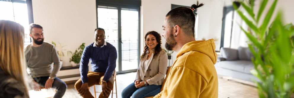 A diverse group of smiling people sit in a circle of chairs facing each other.