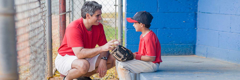 A father crouches in a dugout in front of his son, who wears a baseball uniform and glove. He hands his son a baseball.