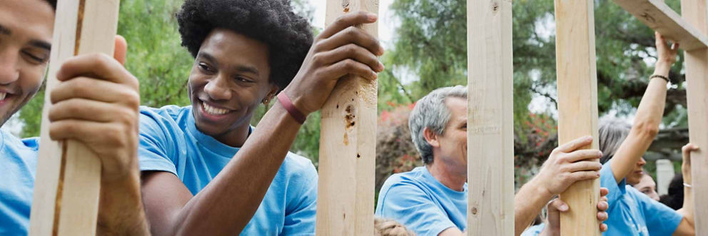Several volunteers of different ages and ethnicities hold up wooden framing as they build a house.