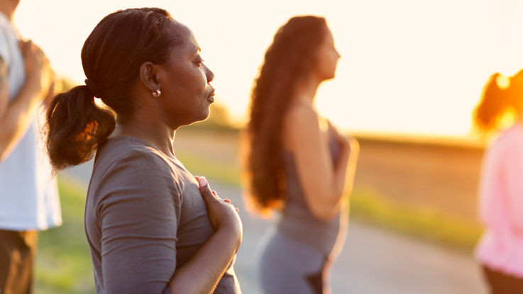 In a field surrounded by her yoga class, a young woman stands in a pose with hands to her chest.