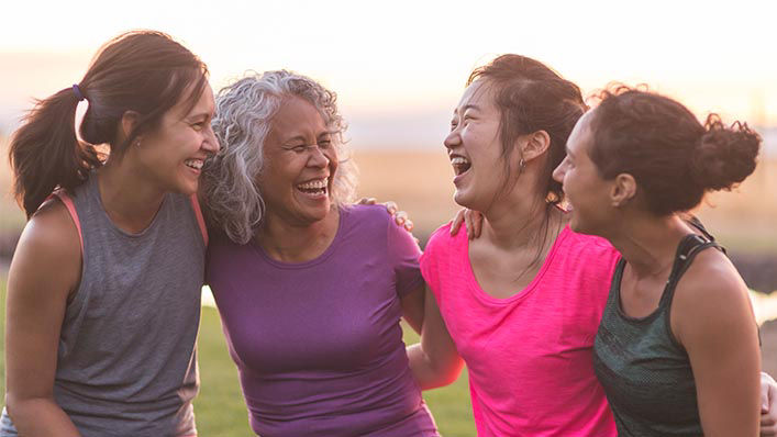 A group of women of different agem laughing and smiling with their arms around each other.