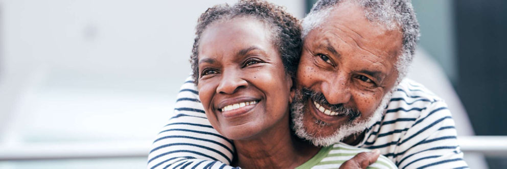 Older man hugs older woman from behind as both smile and look into the distance.