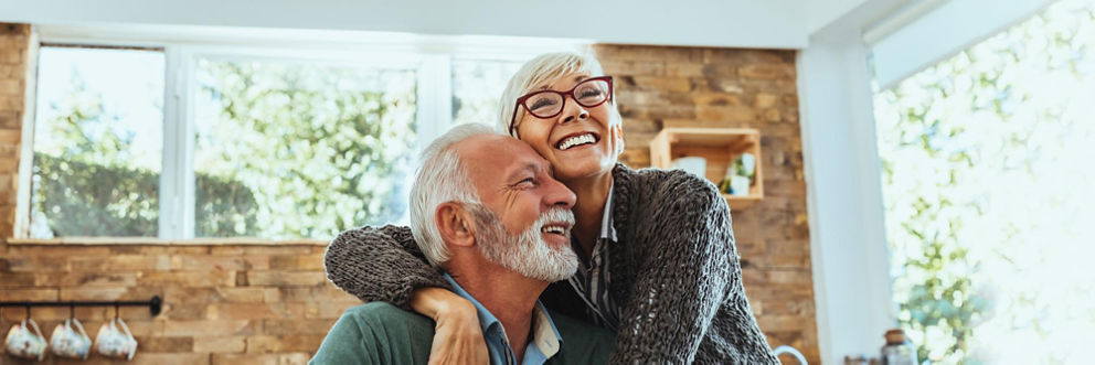An older couple laughs as they embrace in a bright kitchen.