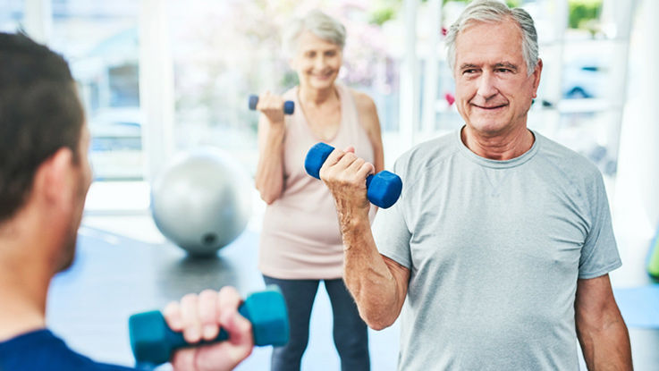 An older man and older woman work out using dumbbells as a personal trainer instructs them