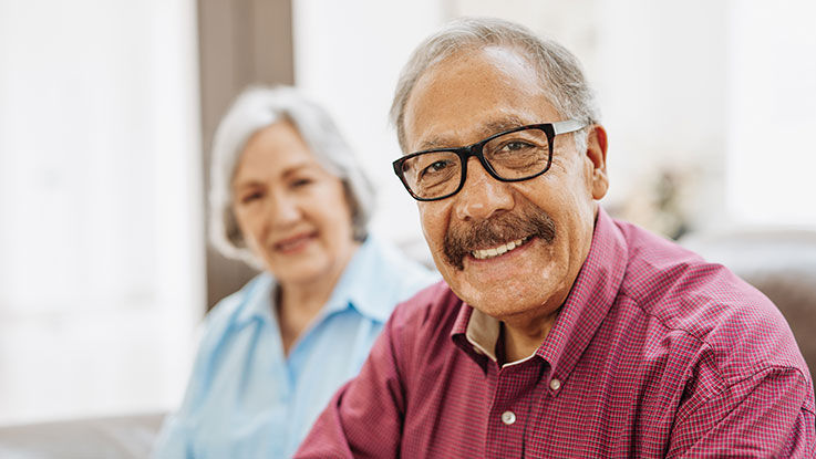 An older man wearing glasses and sitting on a couch smiles and looks at the camera as an older woman sits on the couch behind him