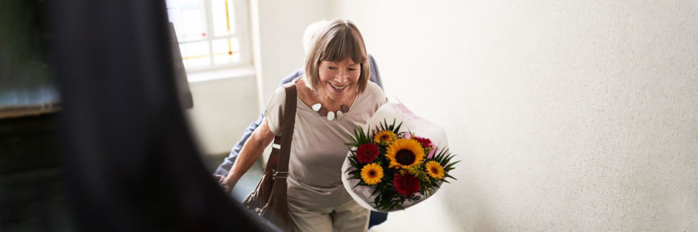 An older woman holding a bouquet of flowers climbs the stairs with her husband close behind.