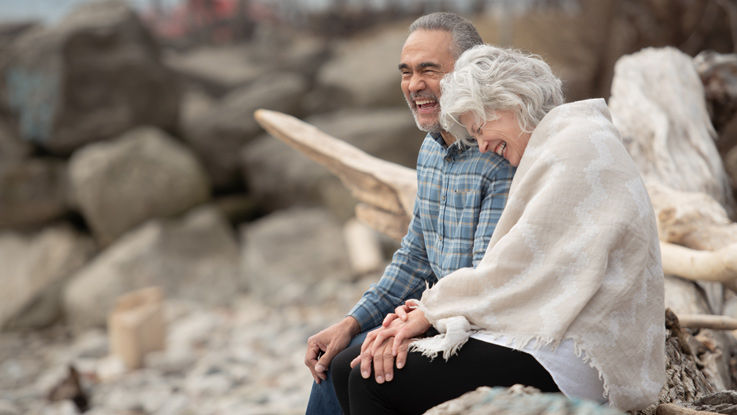 Sitting on a boulder by the beach, an older man and woman laugh as they hold hands.