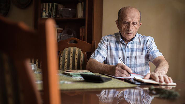 An older man sits at his kitchen table as he fills out forms.