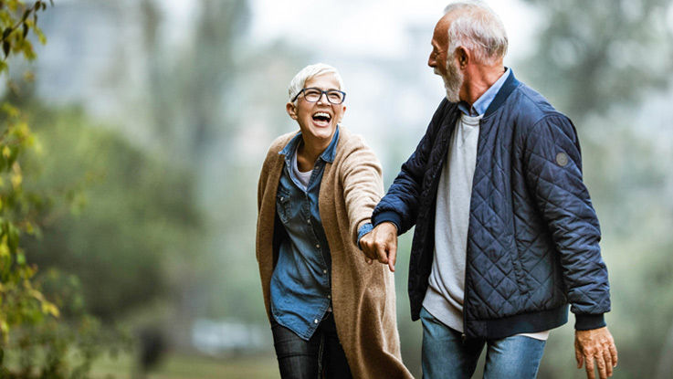 An older couple smiles at each other while holding hands on a walk in the park.