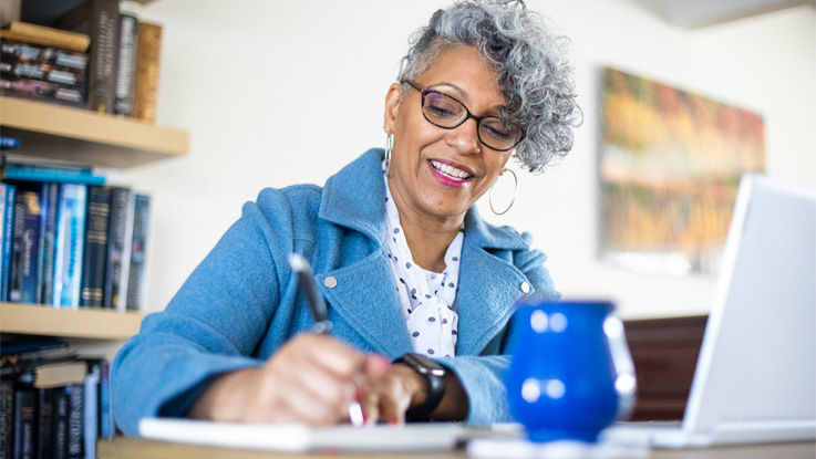 A senior woman sitting at a desk takes notes on a notepad while looking at her Medicare options on a laptop. 